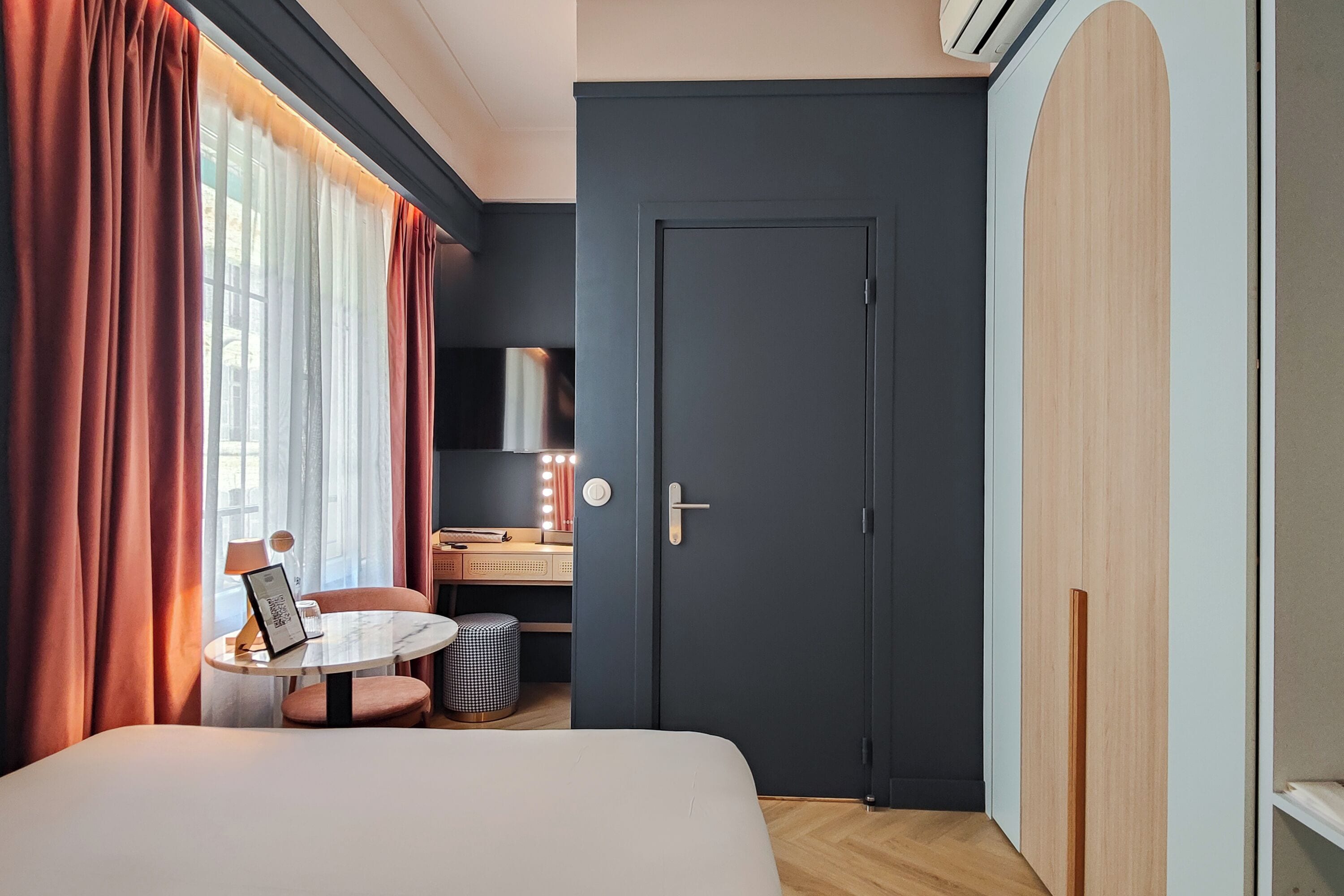 A hotel room with a bed, a round table with a chair, a desk with a chair, a trash bin, and a door.