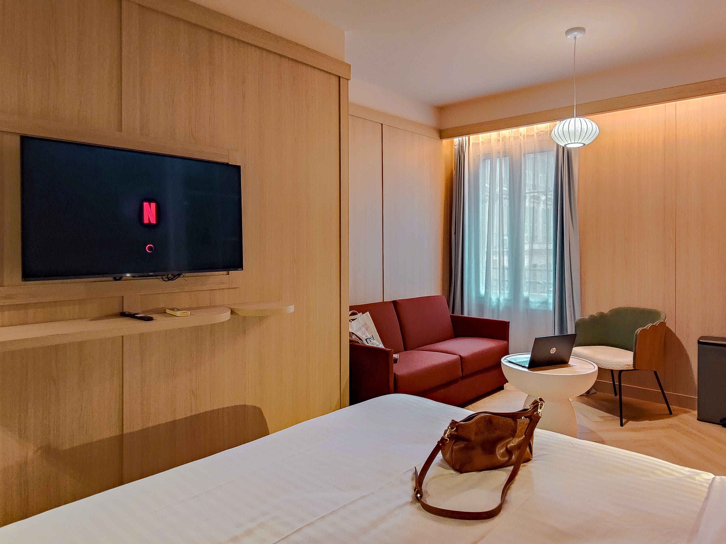 Una camera d'albergo con una TV a schermo piatto che mostra il logo Netflix, un divano rosso, un tavolino con un portatile, un letto bianco e una borsa marrone sul letto.