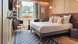 A hotel room with a bed, a desk, a chair, a small table, a lamp, and a view of buildings outside. - Hotel Aparthotel AMMI Nice Lafayette (Nice)