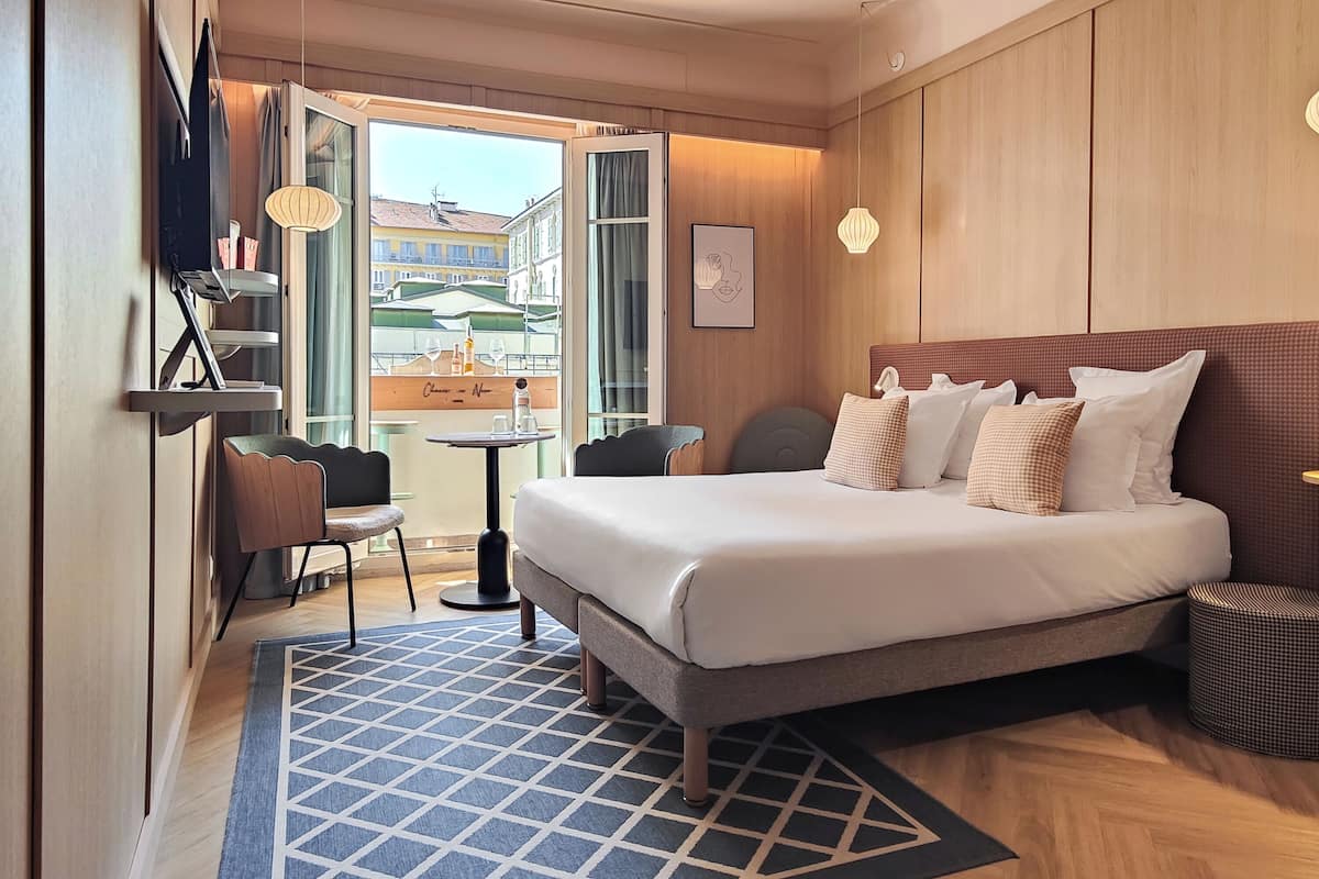 Habitación de hotel con cama, escritorio, silla, una mesita, lámpara y vistas a edificios.