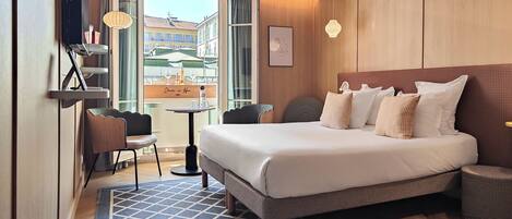 A hotel room with a bed, a desk, a chair, a small table, a lamp, and a view of buildings outside.
