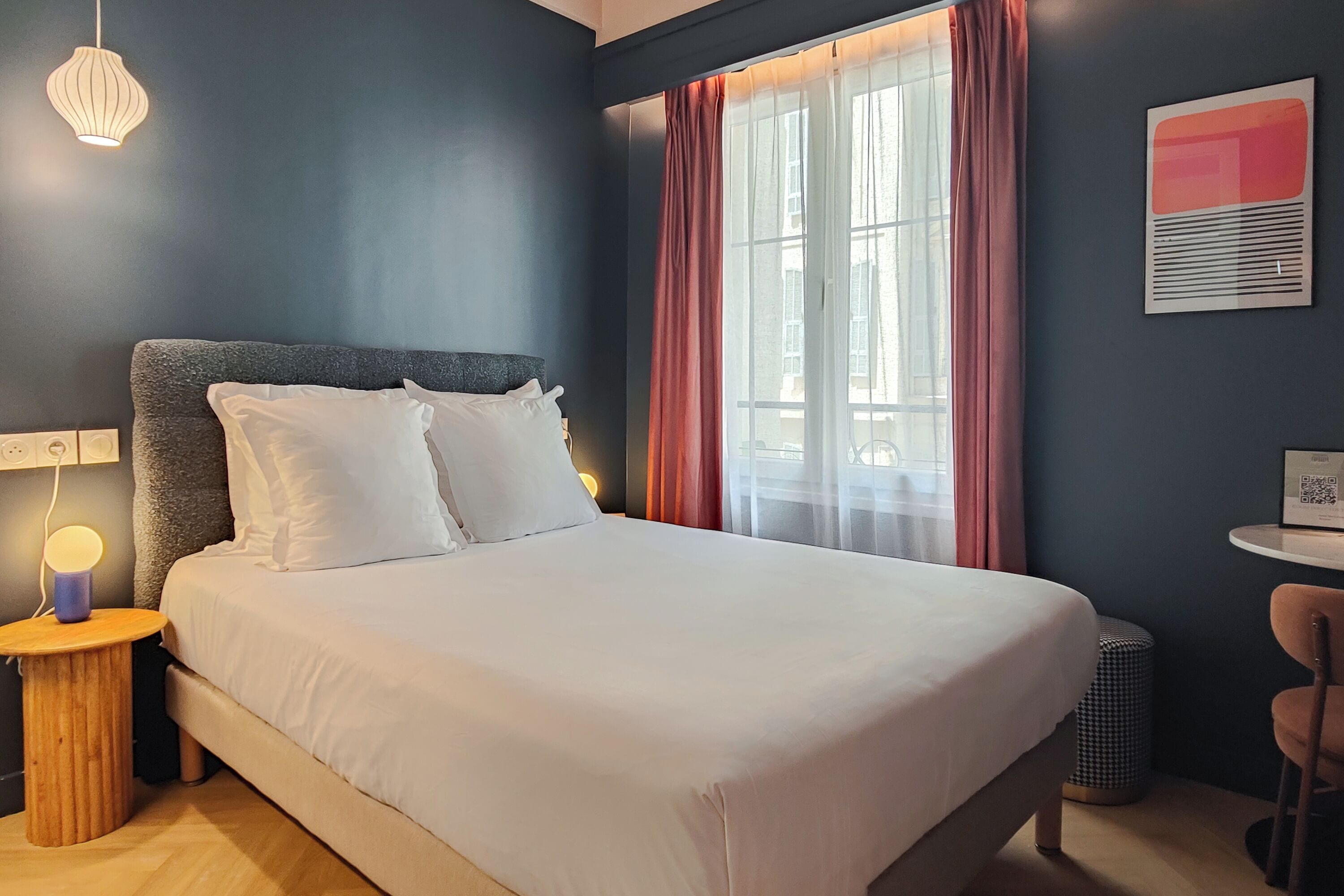 A neatly made bed with white linens, a round wooden nightstand with a lamp, and a window with sheer curtains.