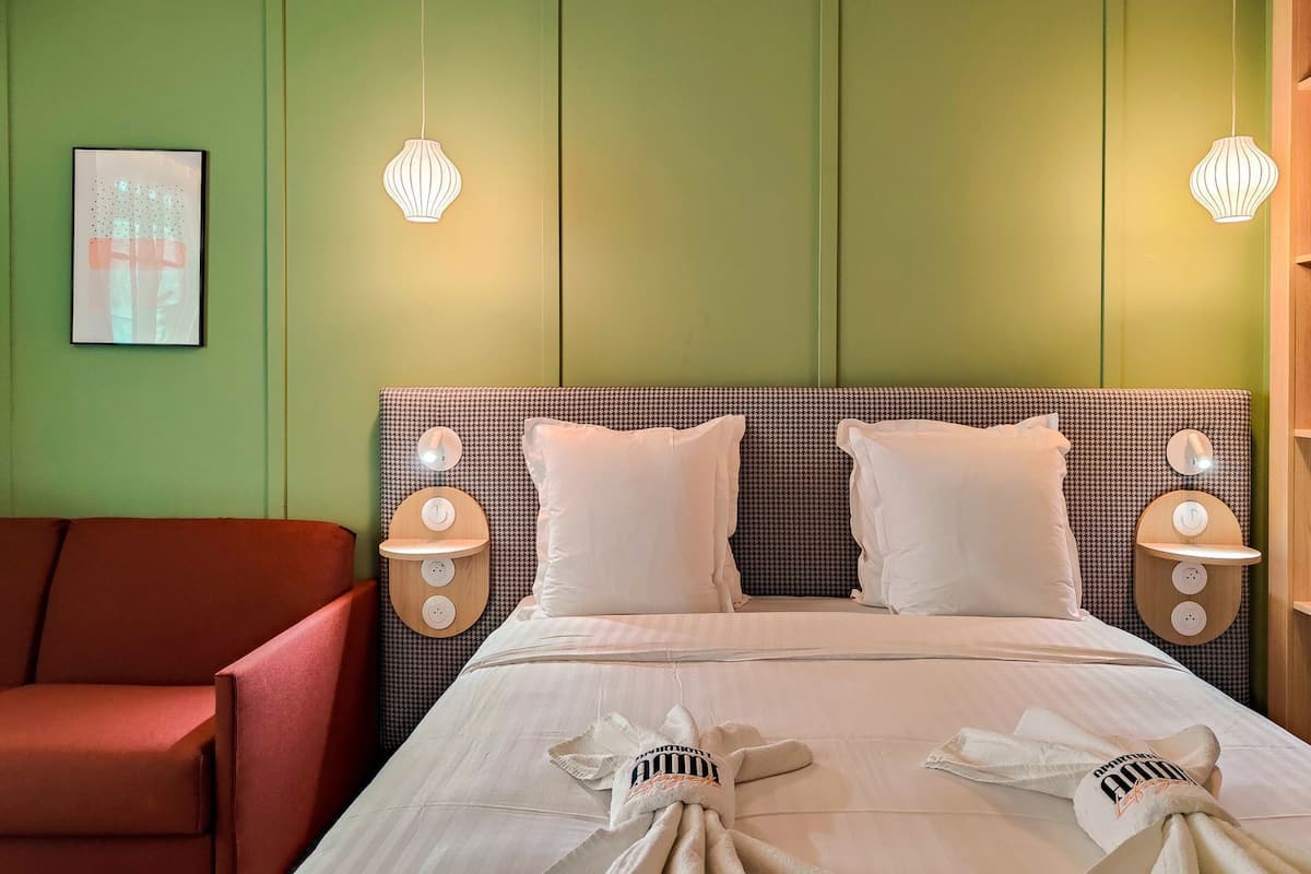 Una habitación de hotel con una cama, dos mesitas de noche con lámparas, un sillón rojo y un cuadro en la pared.