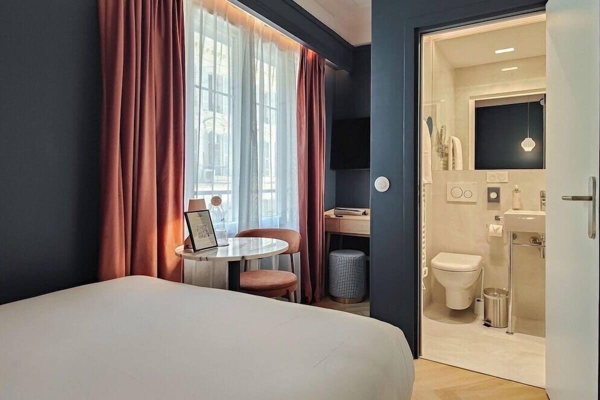A hotel room with a bed, a small table with a chair, a desk with a chair, a window with curtains, and a bathroom with a toilet, sink, and mirror.