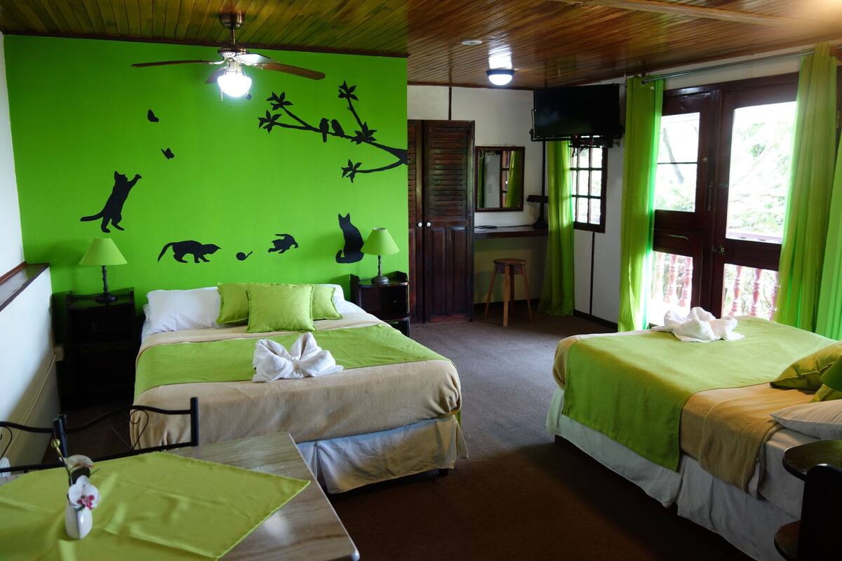 Habitación de hotel con dos camas, un escritorio, una silla, un ventilador de techo y una pared verde con siluetas de animales.