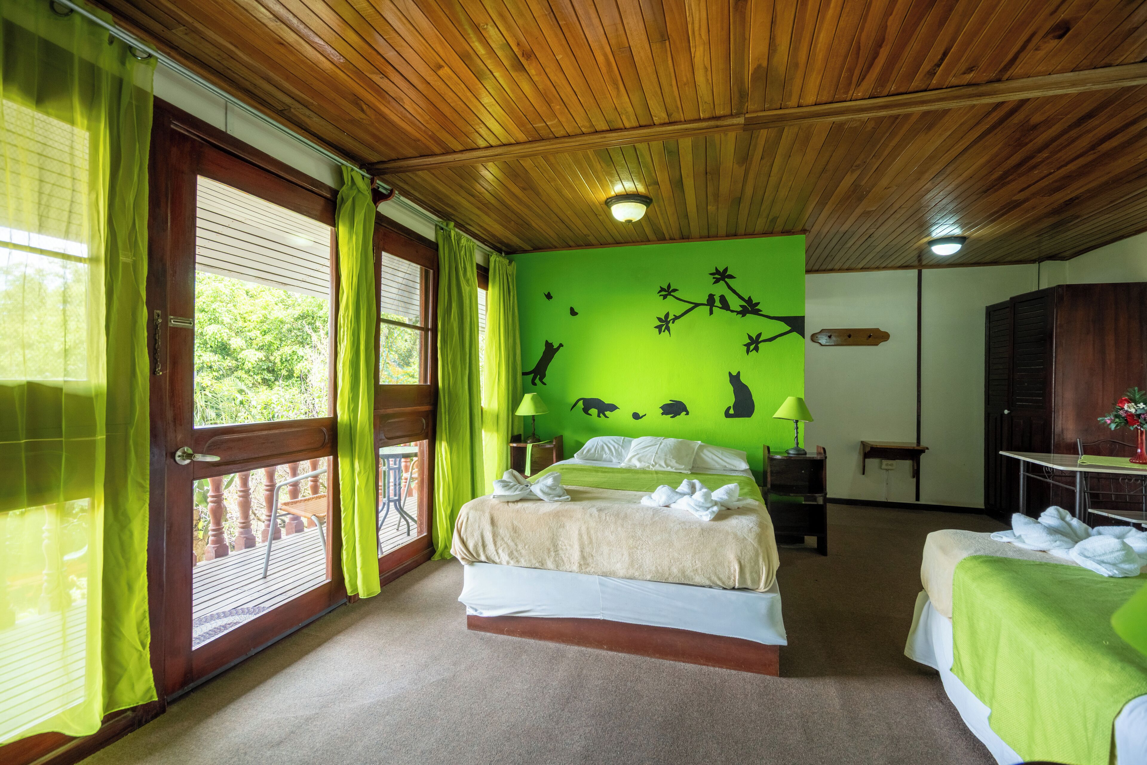 A room with a wooden ceiling, a bed with a green wall mural, a second bed, a nightstand, and a window with curtains.