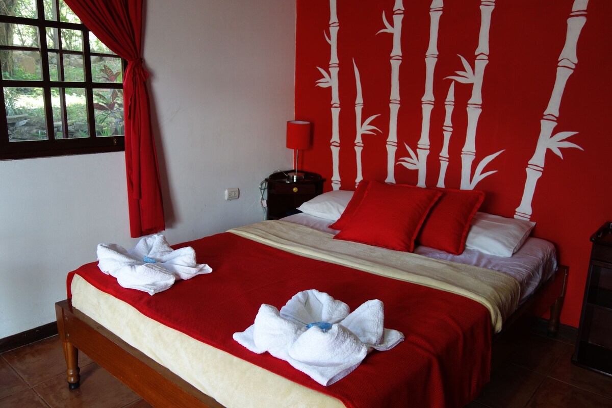 Une chambre d’hôtel avec un lit, une literie rouge et blanche, un rideau rouge, un mur orné de motifs en bambou, une table de chevet avec une lampe et une fenêtre donnant sur un paysage verdoyant.