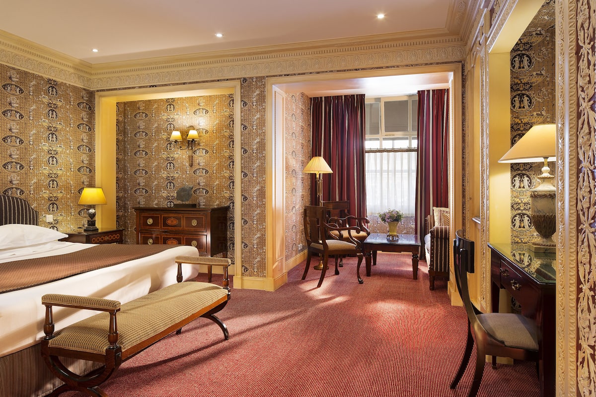 Ein Hotelzimmer mit einem großen Bett, einer Sitzecke mit Sessel und Tisch, einem Schreibtisch mit Lampe und einer Wand mit Musterung.