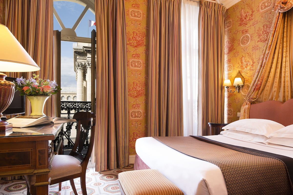 Ein Hotelzimmer mit Bett, Schreibtisch mit Lampe und Blick auf ein Gebäude durch das Fenster.