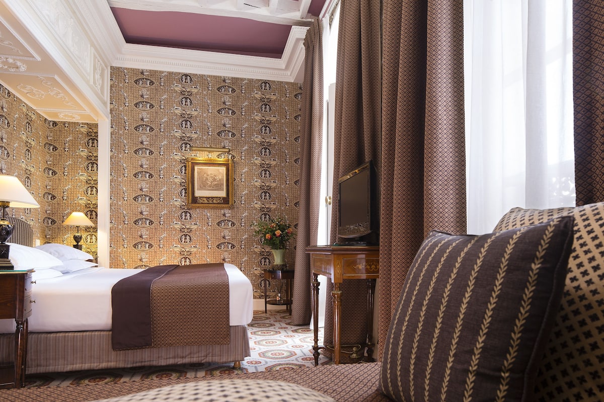 Ein Hotelzimmer mit einem Bett, einem Schreibtisch, einem Stuhl, einem Fernseher und einem Fenster mit Vorhängen.