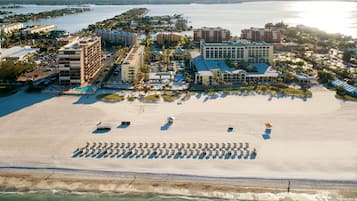 Vista aérea de un complejo turístico costero con una extensa playa de arena, varios edificios de hotel y un puerto deportivo.