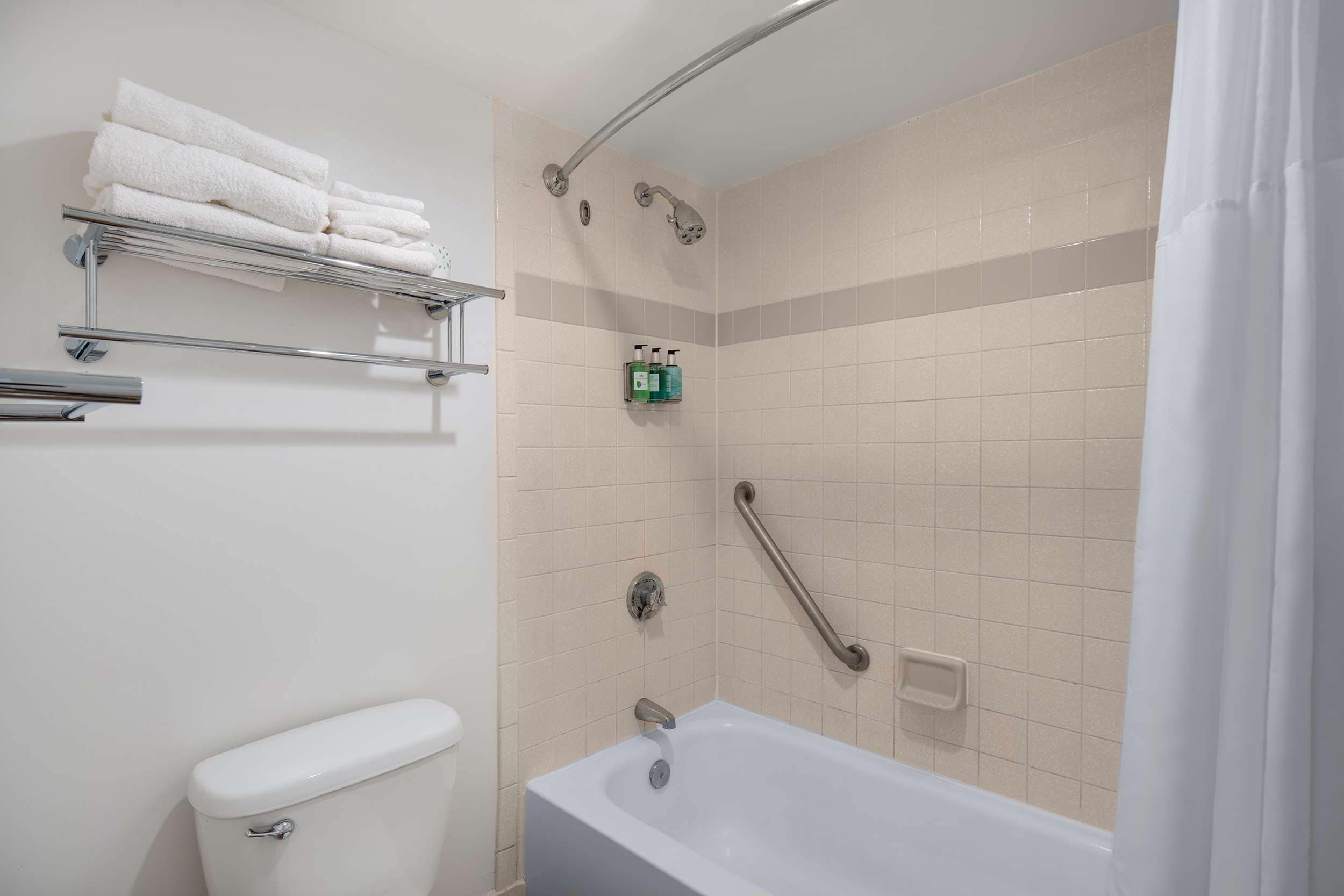 A bathroom with a white bathtub, a toilet, a towel rack with folded towels, a shower with a handheld showerhead, and a grab bar.