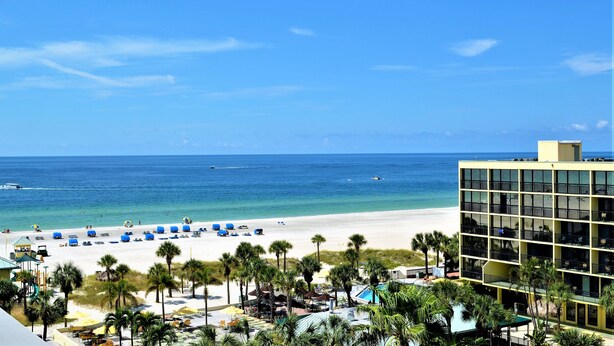A beach view with a hotel building, palm trees, and beach umbrellas. - St Pete Shores Hotel (St. Pete Beach)