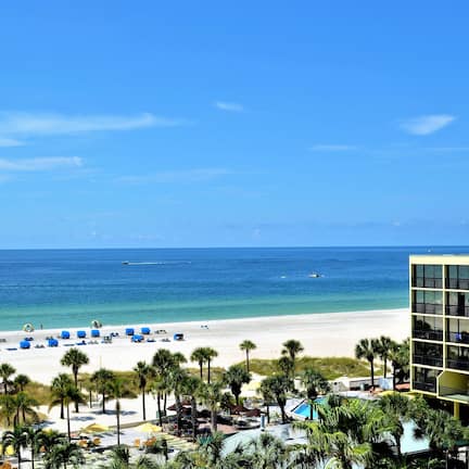 A beach view with a hotel building, palm trees, and beach umbrellas.