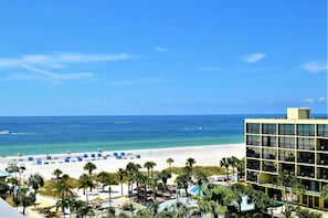 Vista de una playa con un edificio de hotel, palmeras y sombrillas.