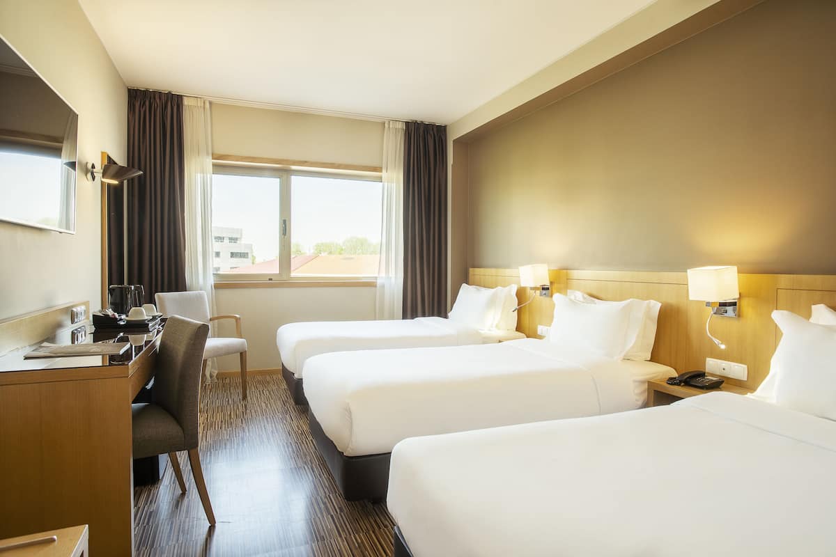 A hotel room with three beds, a desk with a chair, a lamp, and a window with curtains.