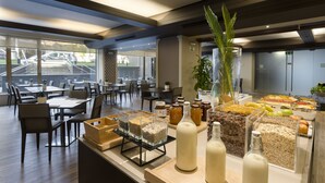 A breakfast buffet with various food items and beverages, a seating area with tables and chairs, and a view of the outside through large windows. - HF Tuela Porto (Porto)