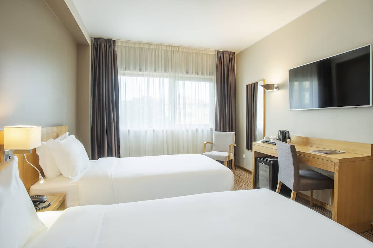 A hotel room with two beds, a desk, a chair, a TV, and a window with curtains.