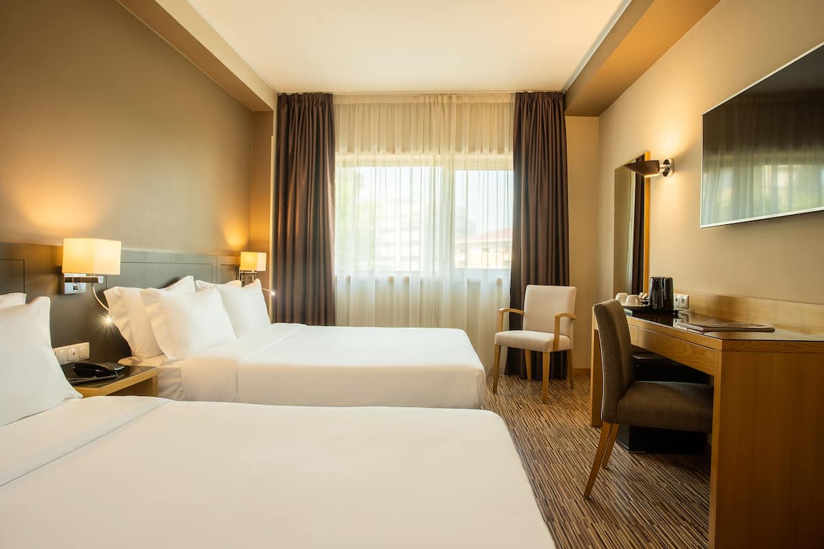 A hotel room with two beds, a desk, a chair, a TV, and a window with curtains.