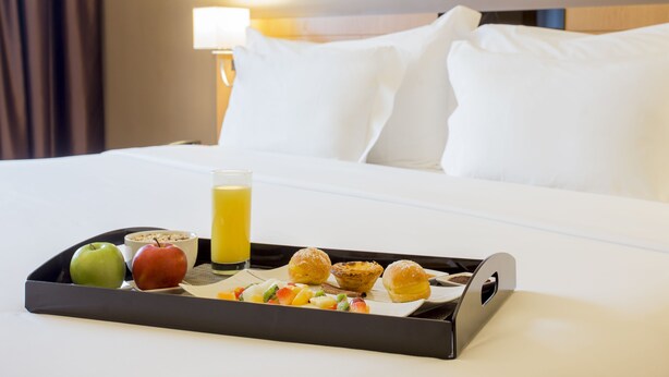 A breakfast tray with fruits, pastries, and a glass of orange juice on a bed in a hotel room. - HF Tuela Porto (Porto)