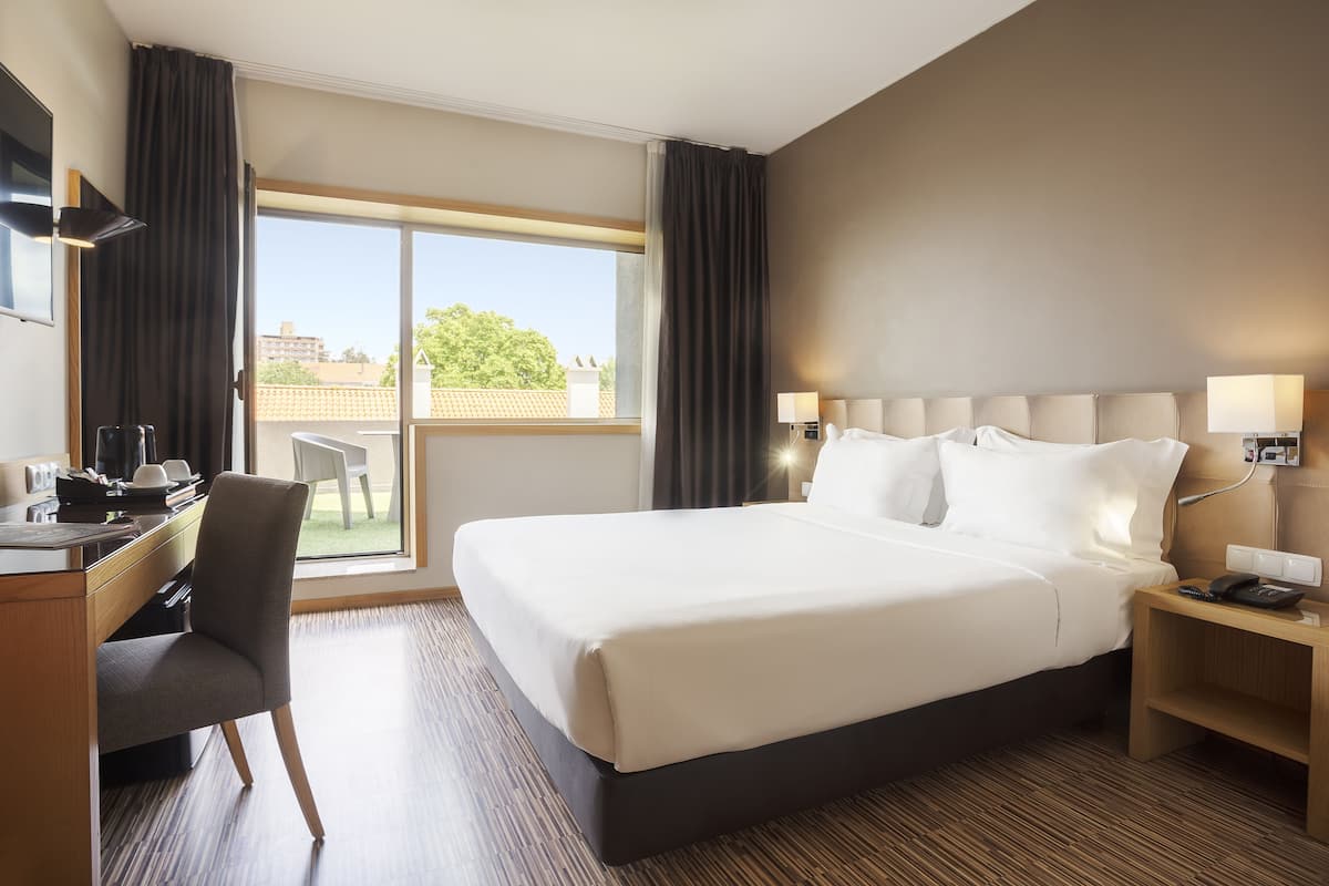 A hotel room with a large bed, a desk, a chair, and a view of the outdoors.