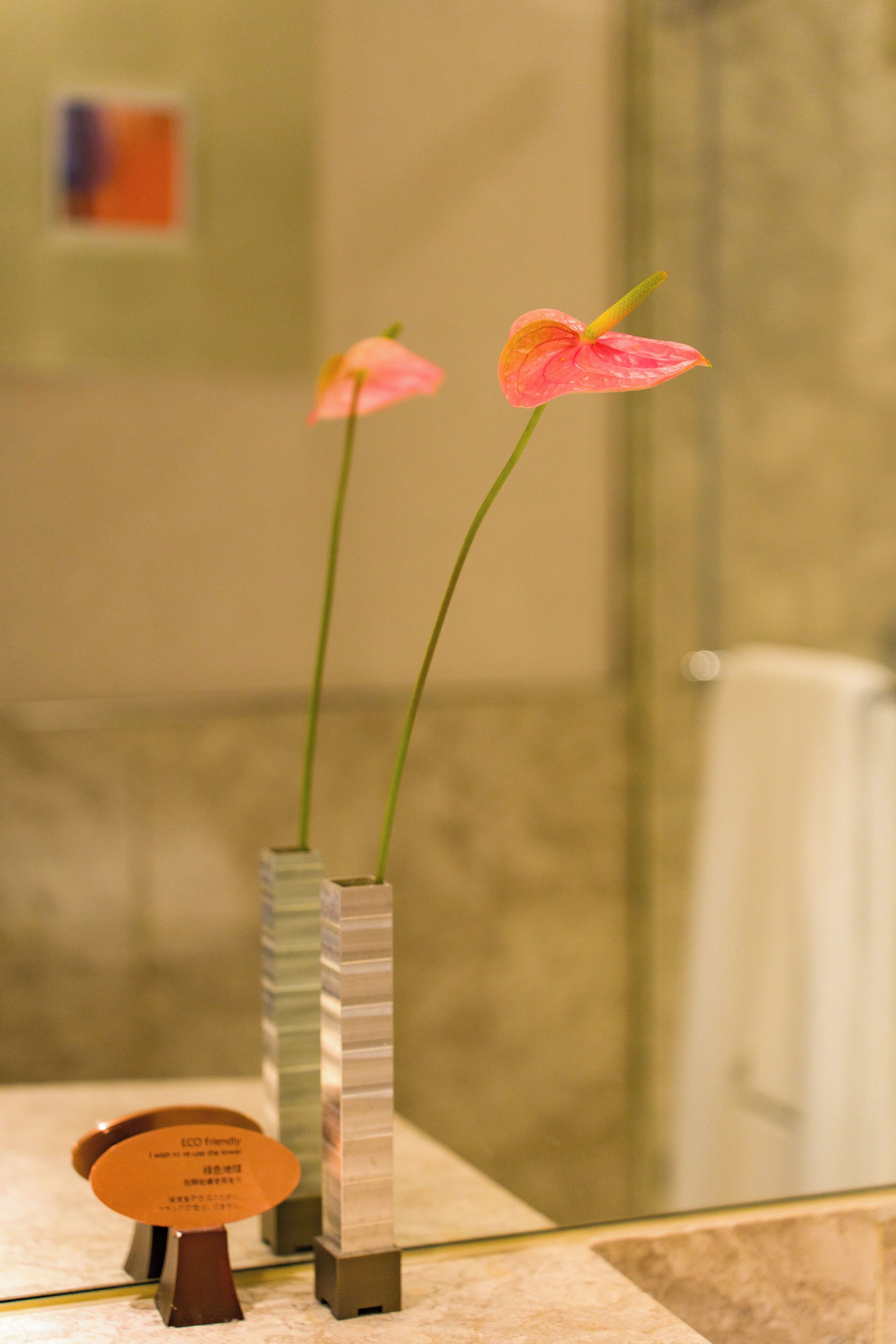 Ein Badezimmer mit zwei Glasvasen mit rosa Blumen, einer Tafel mit Text und einem Spiegel, der den Raum widerspiegelt.