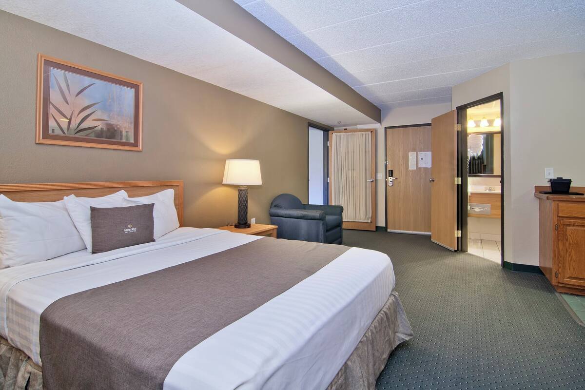 Een hotelkamer met een groot bed, een bureau met lamp, een stoel, een houten garderobe, en een zichtbare badkamer door een geopende deur.