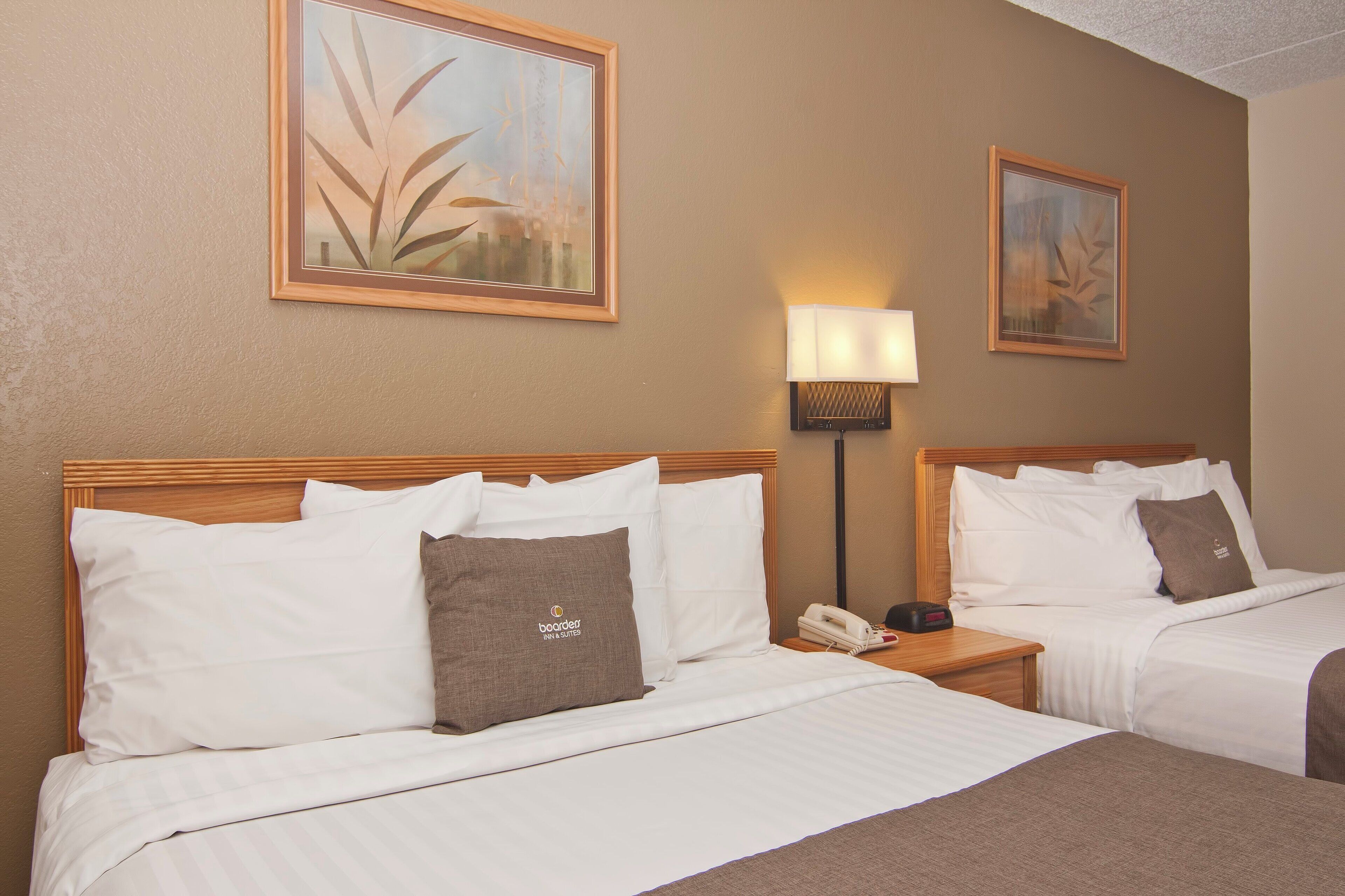 Habitación de hotel con dos camas, cada una con ropa blanca y almohadas marrones, una mesita de noche con lámpara y cuadros en las paredes.