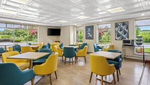 A modern dining area with yellow and teal chairs, wooden tables, and a large window offering a view of greenery. - Comfort Inn & Suites Walterboro I-95 (Walterboro)