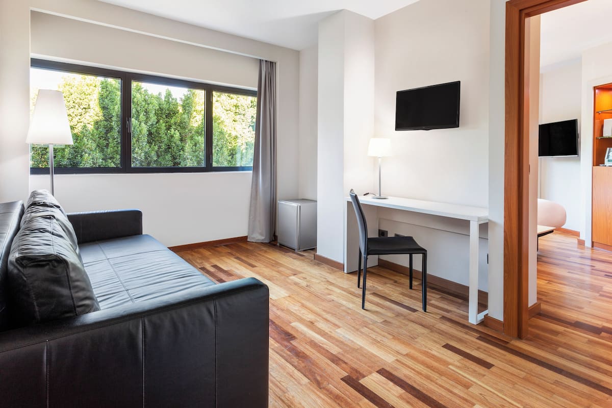 Una sala de estar moderna con un sofá, un escritorio y una silla. Hay una ventana con vistas a árboles, un televisor de pantalla plana montado en la pared y una lámpara en el escritorio.