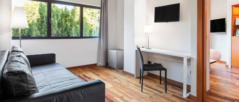 A modern living room with a sofa, a desk, and a chair. There is a window with a view of trees, a flat-screen TV mounted on the wall, and a lamp on the desk.