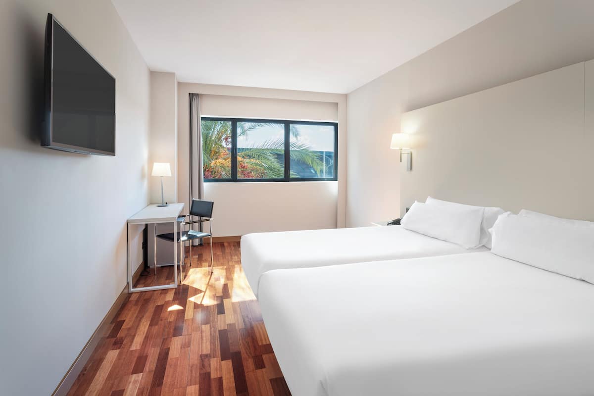 Habitación de hotel con dos camas, un escritorio, una silla, un televisor y una ventana con vistas a palmeras y al océano.