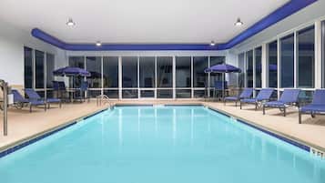 An indoor swimming pool with clear blue water, surrounded by lounge chairs and umbrellas, and large windows providing natural light.