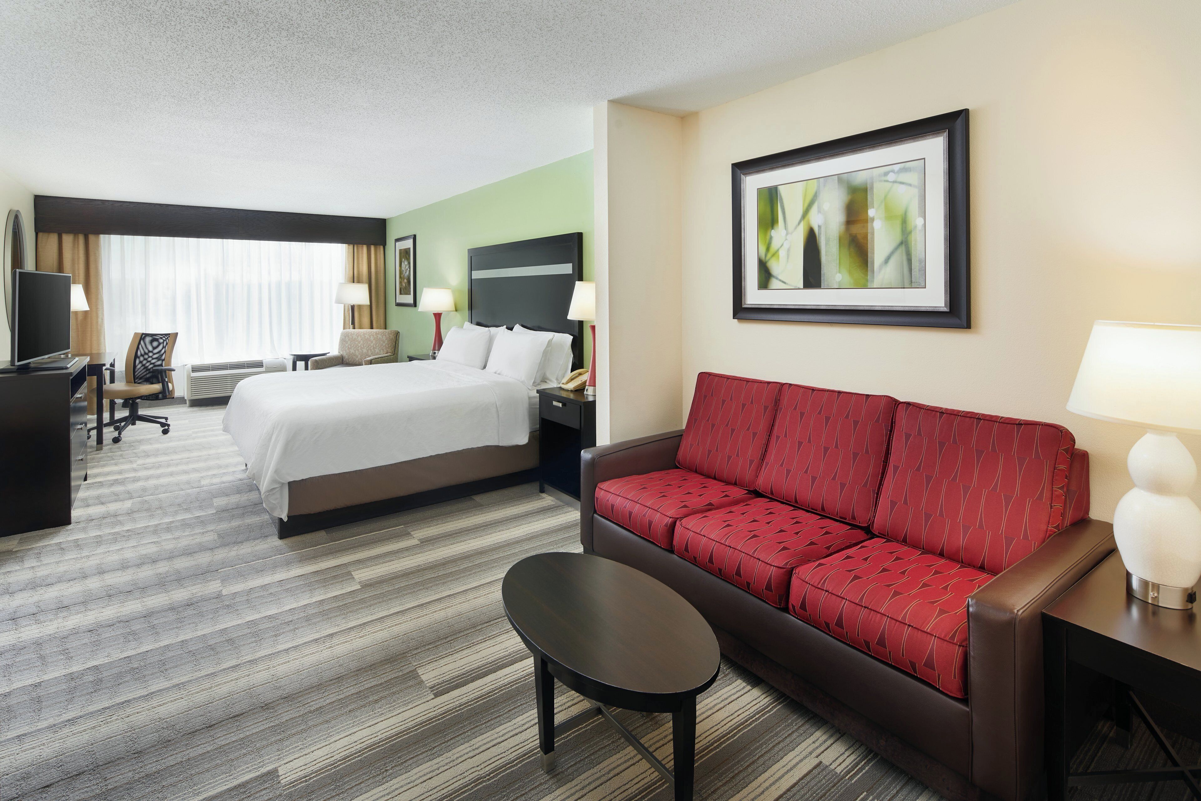 A hotel room with a bed, a red sofa, a small table, a TV, and a framed picture on the wall.