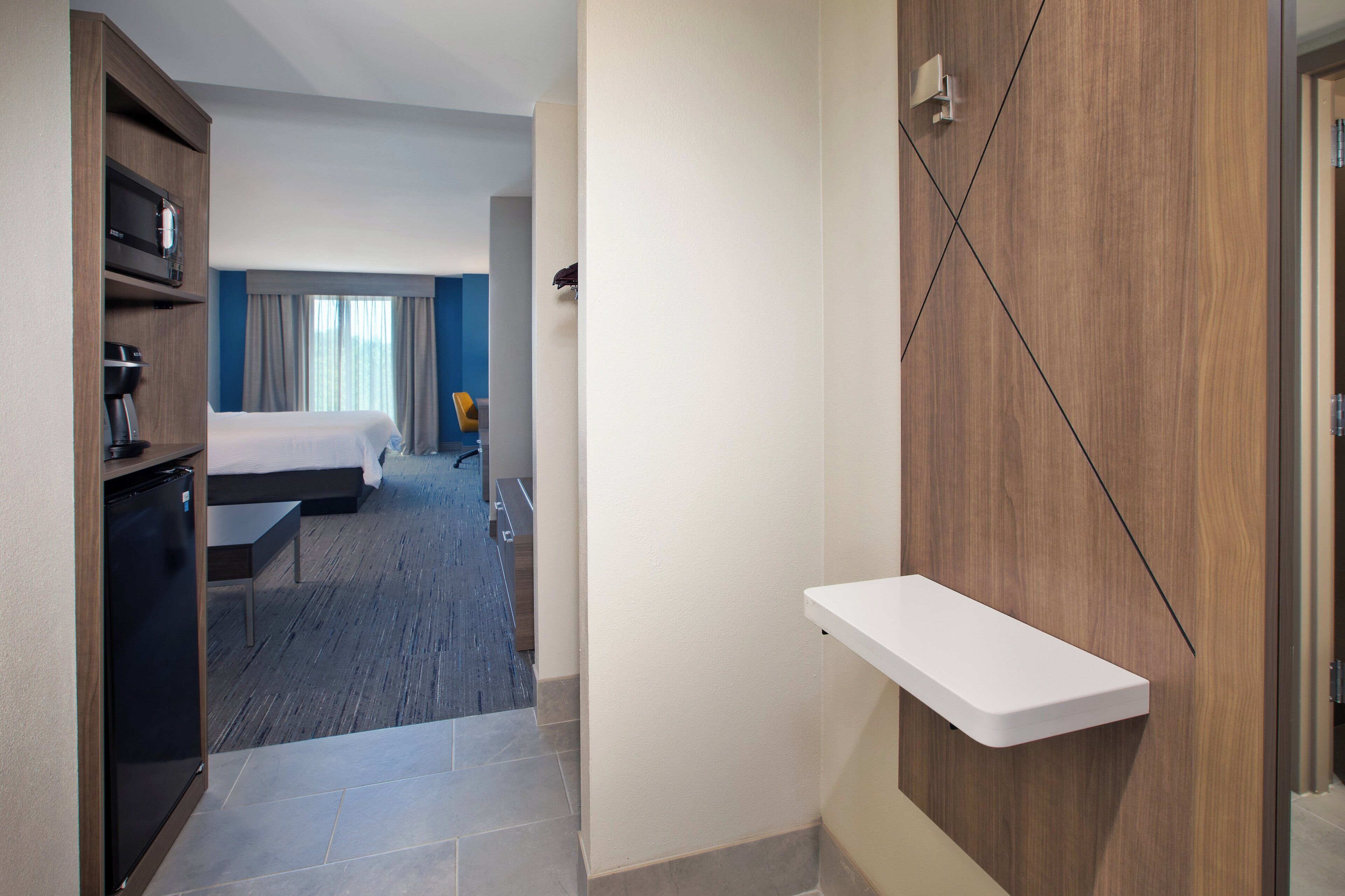 Hotellihuone, jossa on sänky, työpöytä, kahvinkeitin ja pieni valkoinen hylly.