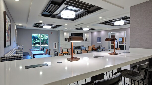 Een moderne hotel lobby met een lange receptiebalie, een bar met krukken, zitplaatsen met patroonkussens en een groot raam met uitzicht op groen.