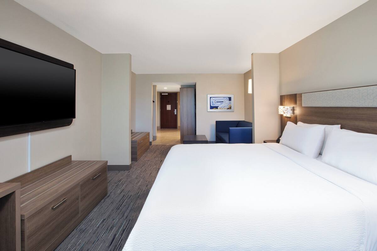 Une chambre d’hôtel dotée d’un grand lit, d’un téléviseur à écran plat fixé au mur, d’un bureau avec des tiroirs et d’un petit coin salon comprenant une chaise et une table.