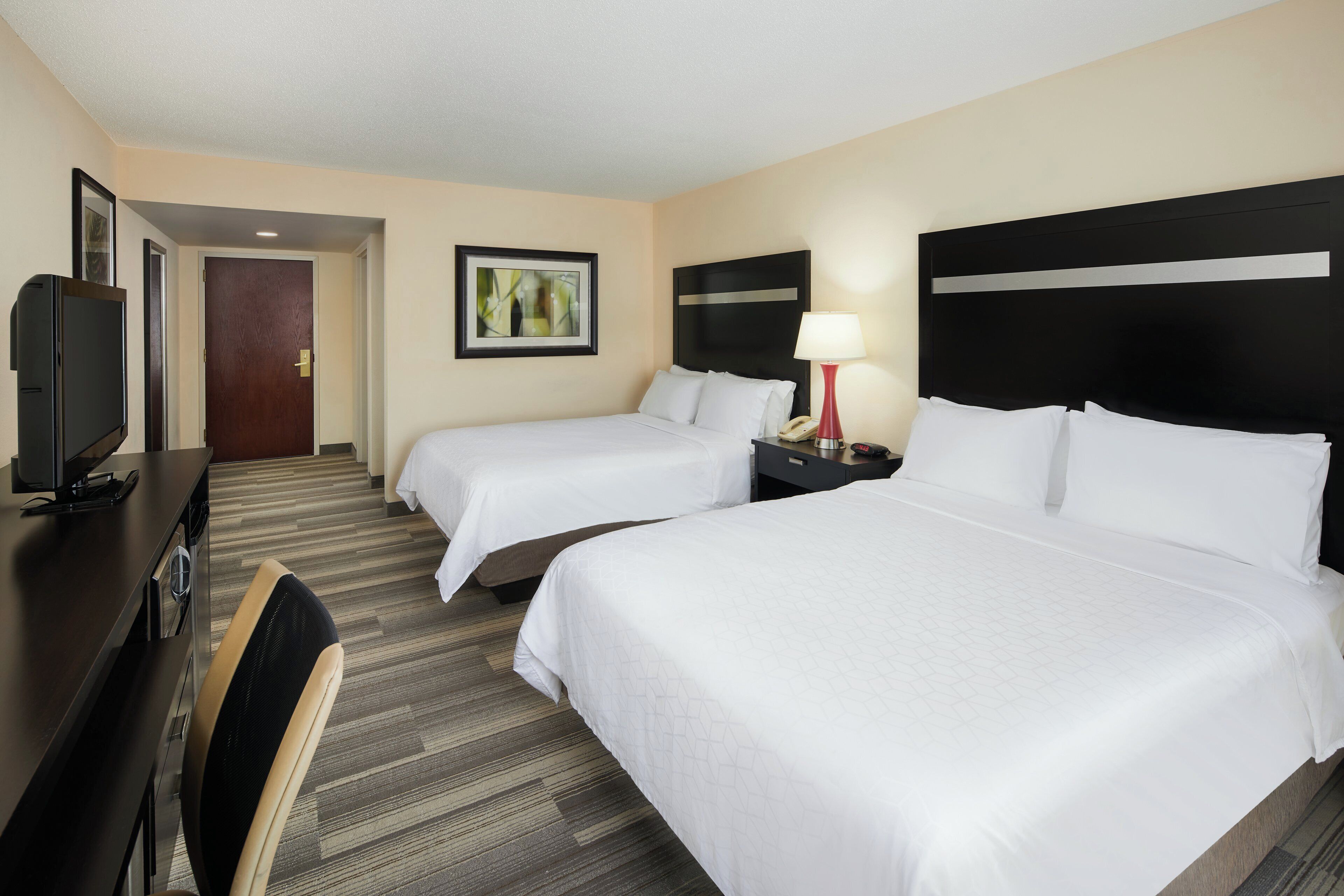 Quarto de hotel com duas camas, uma escrivaninha com televisão, um quadro na parede e um abajur na mesa de cabeceira.