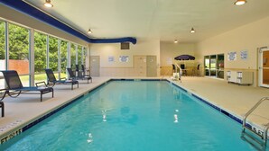 Ein Hallenbad mit klarem Wasser, einem blauen Rand und einem Rettungsschwimmerstand. Liegestühle und eine überdachte Sitzgelegenheit sind vorhanden.