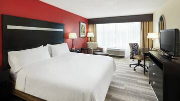 Quarto de hotel com uma cama grande, escrivaninha com computador, cadeira, televisão e janela com cortinas.
