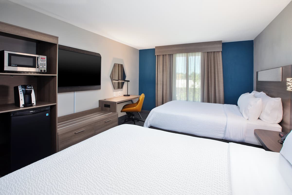 Ett hotellrum med två sängar, ett skrivbord med en stol, en mikrovågsugn, ett kylskåp och en platt-TV.