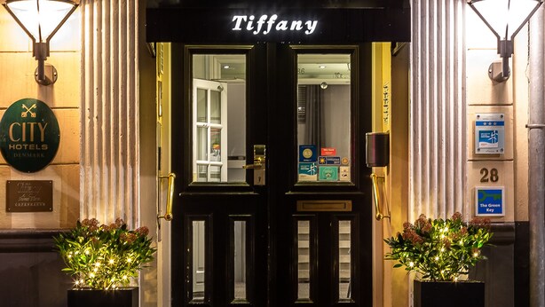 Exterior de um edifício com fachada preta e o nome 'Tiffany' acima da entrada. Há duas grandes plantas em vasos na entrada, e uma placa indicando que o edifício faz parte da rede 'CITY HOTELS DENMARK'.