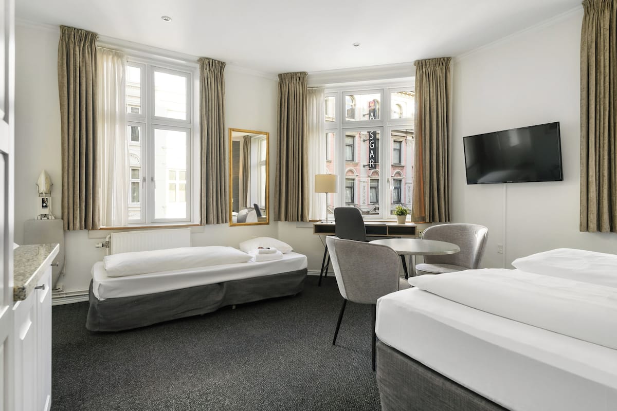 Habitación de hotel con dos camas, un escritorio, una silla, un televisor y ventanas grandes con cortinas.