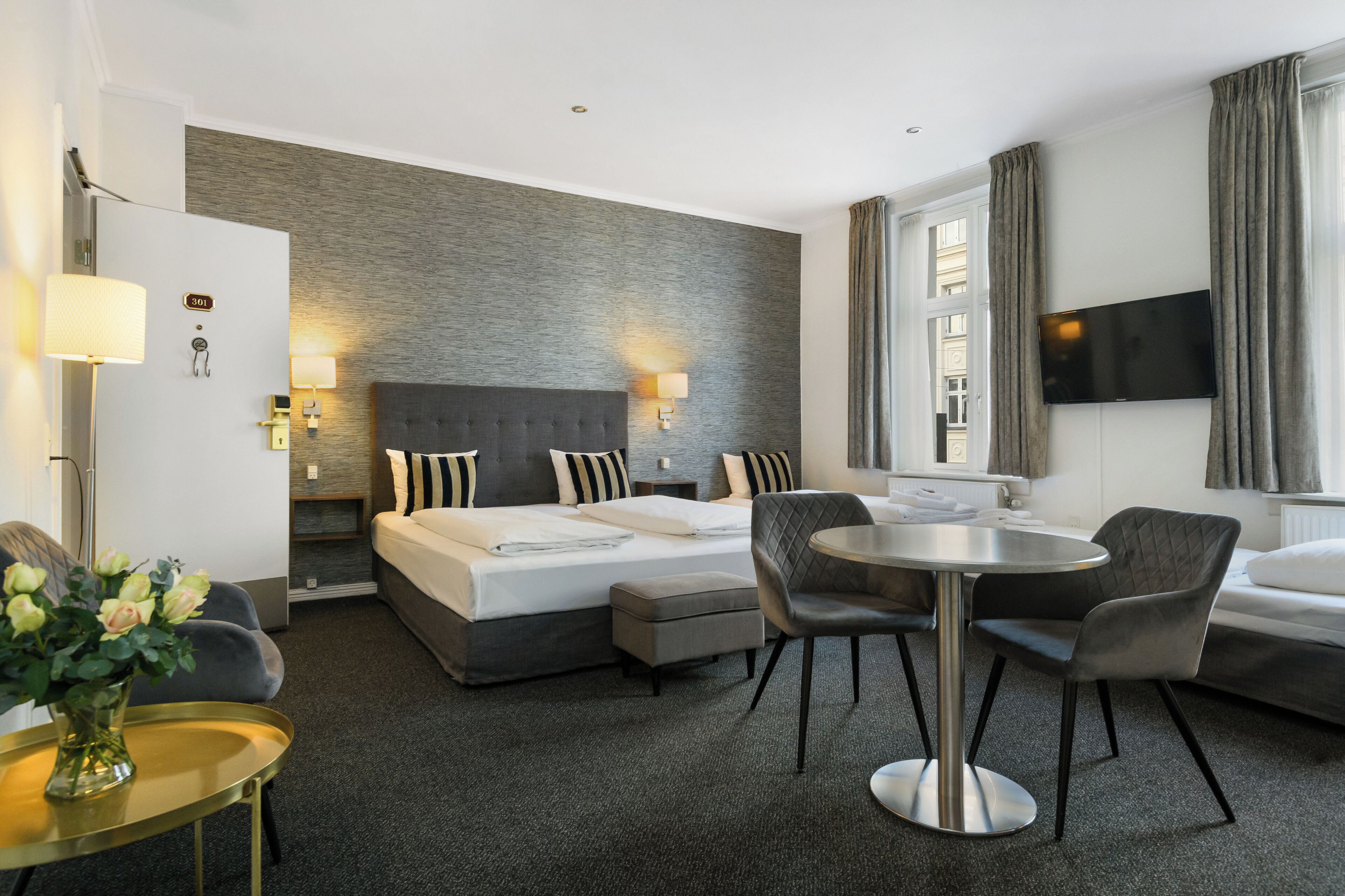 A modern hotel room with a large bed, a round dining table, two chairs, a TV, and a small side table with a vase of flowers.