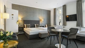A modern hotel room with a large bed, a round dining table, two chairs, a TV, and a small side table with a vase of flowers.