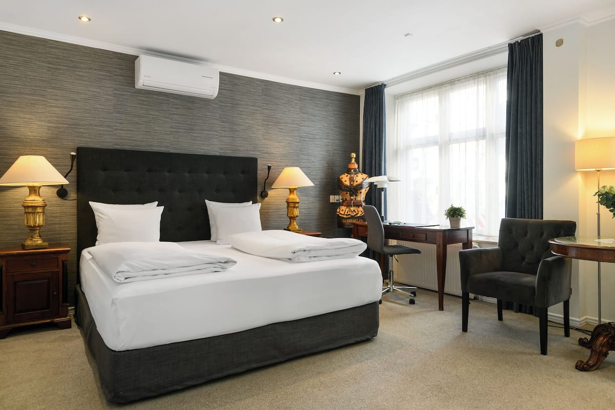Habitación de hotel con una cama grande, dos mesitas de noche con lámparas, un escritorio, una silla y una ventana con cortinas.