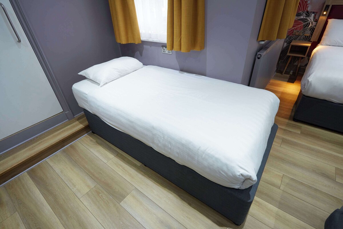 A single bed with white bedding in a room with wooden flooring and a window with yellow curtains.