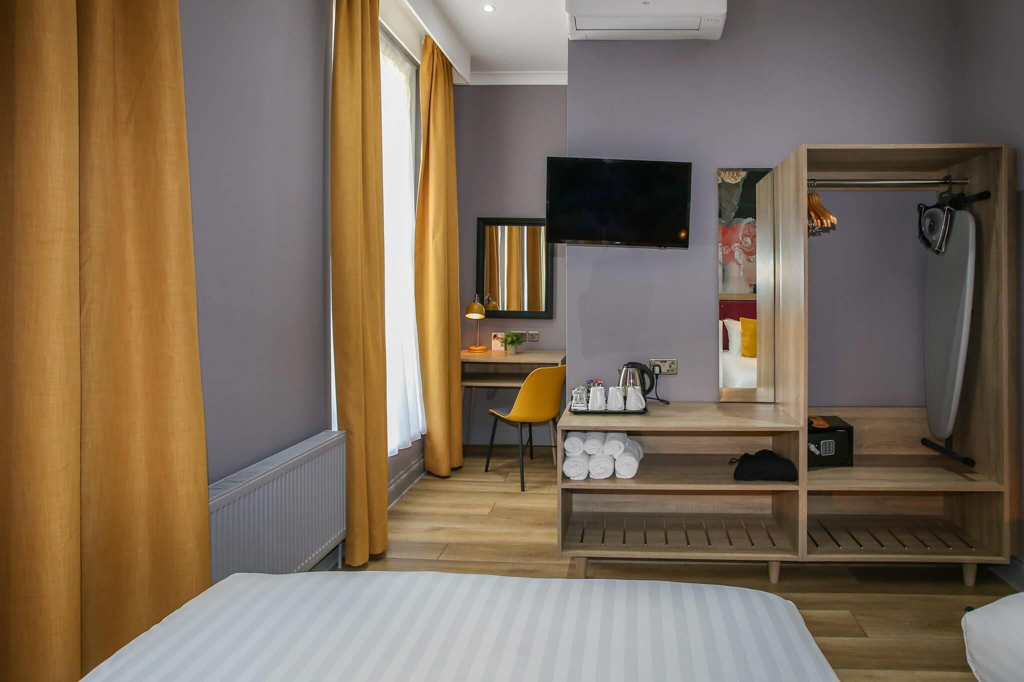 Quarto de hotel com cama, secretária e cadeira, televisão, guarda-roupa e prateleira com produtos de higiene pessoal.