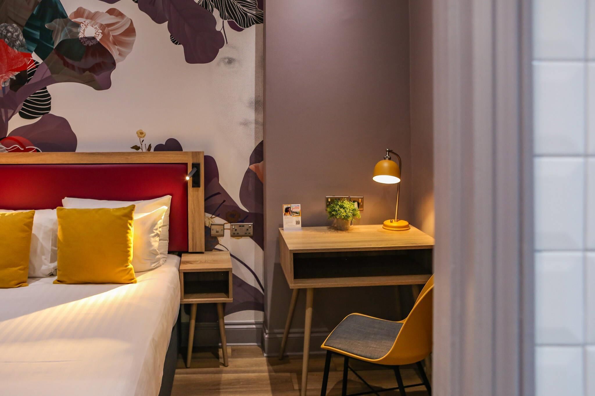 A hotel room with a bed, a desk, a chair, a lamp, and a wall with a floral mural.