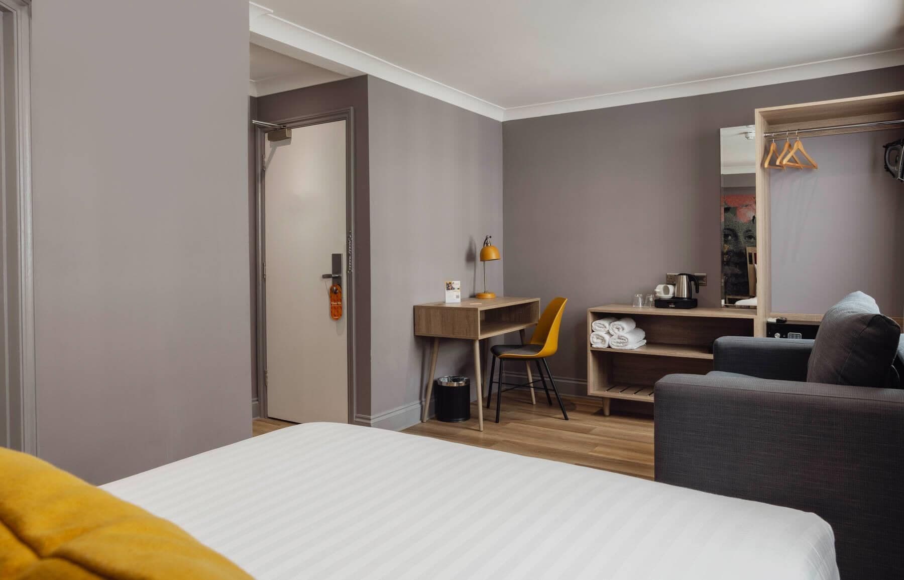 A hotel room with a bed, a desk with a chair, a wardrobe, and a mirror.